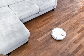 Robotic vacuum cleaner runs near sofa in room on laminate floor. Robot controlled by voice commands...