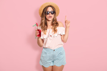 Attractive young woman in summer outfit isolated on pink background. Happy girl in sunglasses posing with straw hat and cocktail, spending hot summer days on beach, points fore finger up, has new idea