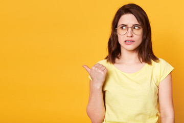 Good looking brunette female dressed in casual yellow t shirt and stylish eyeglasses, looking aside, showing direction with her thumb, looks astonished. Emotions concept. Copy space for advertisement.