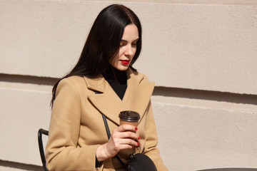 Charming young brunette woman with long dark hair wearing coat, sitting at table in street cafe and drinking coffee or tea, attractive female has thoughtful facial expression, worried about her work.