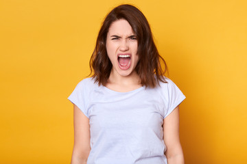 Obraz premium Screaming emotional angry woman isolated over yellow studio background. Half length portrait of young brunette female wearing white casual t shirt. Human emotions and facial expression concept.