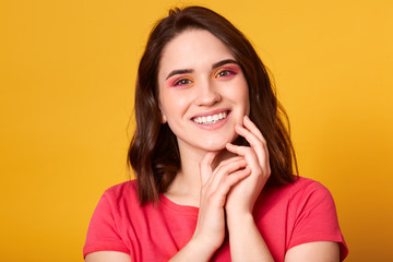 Indoor shot of beautiful female with natural perfect skin and bright make up in pink and orange colours, looking smiling at camera, keeps hands together near face, dressed red t shirt. Beauty concept.