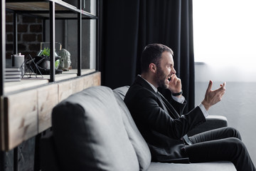 angry man sitting on sofa and quarreling while talking on smartphone in office