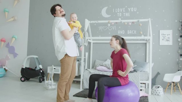 Sporty woman doing exercises sitting on fitness ball while fastidious father giving her crap toddler daughter to change diaper. Young mother training on fitball at home while infant crap one's pants.