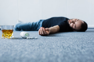 selective focus of adult unconscious man lying on grey floor near containers with pills and glass of whiskey