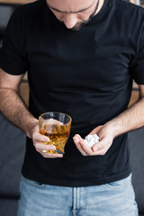 cropped view of depressed man in black t-shirt with handful and pills and glass of whiskey
