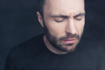 portrait of depressed, handsome bearded man standing by window with closed eyes