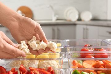 Woman putting cut cauliflower into box and containers with raw vegetables in kitchen, closeup