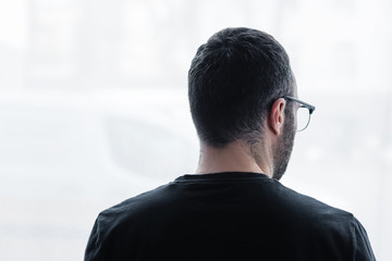back view of adult depressive man in black t-shirt looking out window at home