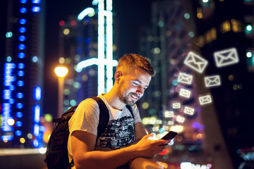 Smiling Caucasian man leaning on railing and sending messages over smart phone. City at night exterior.