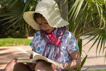 Dziewczynka w słomkowym kapeluszu czyta książkę w cieniu palmy - obrazy, fototapety, plakaty