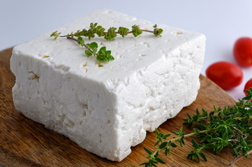Large piece of traditional greek feta cheese on wooden cutting board with oregano and tomatoes on white background