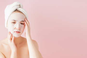 Beautiful young woman is applying a cosmetic tissue mask on a face on a pink background. Healthcare and beauty treatment and technology concept