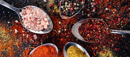 Spoons with different indian spices scattered on background table, peppercorn paprika salt turmeric powder, red, yellow and pink herbs seasoning top view, horizontal banner header for website design
