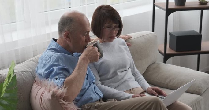 Old couple drinking coffee and using laptop on sofa