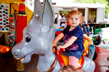 Adorable little toddler girl riding on animal on roundabout carousel in amusement park. Happy...