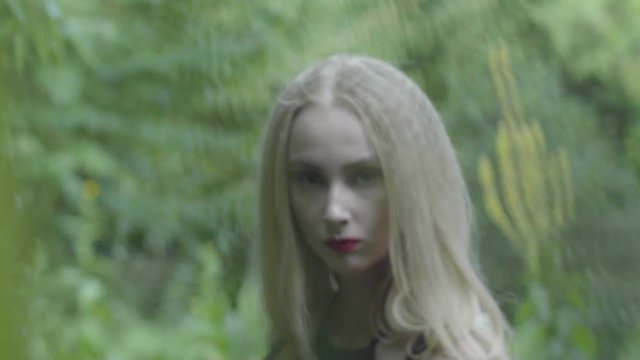 Cute Russian blonde girl posing in a park looking at the camera through the leaves in slow motion 4K. Blurred background
