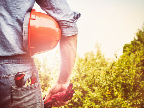 Handsome man in working clothes with tools, standing against a background of trees, blue sky and the rays of the setting sun. View from the back, close-up. Concept of work and employment