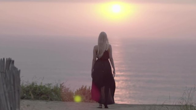 Attractive blond woman posing from behing at sunset, on top of a mountain in wide shot, with grass on the ground in the late afternoon. Beauty shot golden hour 4 K in slow motion.