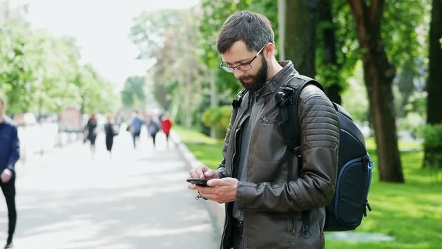 Bearded man with bag uses mobile phone on the street, surf the internet, playing a game, blurred people in the background
