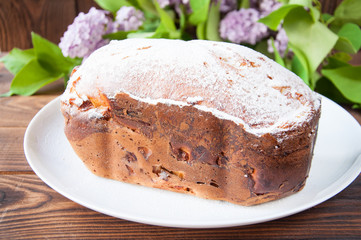 baked homemade cake dusted with powder stands on a white plate near the lilac flowers. On a wooden background.