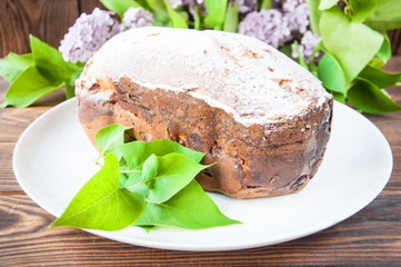 baked homemade cake dusted with powder stands on a white plate near the lilac flowers. On a wooden background.