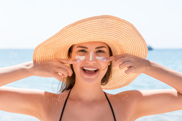 Beautiful young woman in hat is applying sunblock under her eyes and on her nose like Indian. Sun...