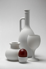 White still life from the stand for the egg with a red egg, a vase, a glass for wine and a jug for wine.