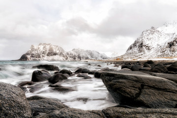 Ocean waves hit the beach With black stones at lofoten and behind the mountains On cloudy days, rain is going to fall in the evening of winter in norway.