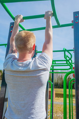 Fototapeta premium The concept of a healthy lifestyle, sports in nature, physical culture. A blond man pulls himself up on a crossbar on a street stadium. Summer sunny day.