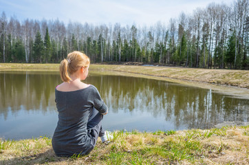 Fototapeta na wymiar The concept of summer outdoor recreation, harmony with nature, trips to the countryside. A young brown-haired girl sits on the shore of a small lake near the forest. Summer sunny day. Copy space.