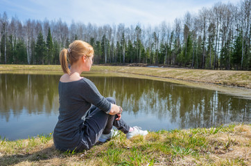 Fototapeta na wymiar The concept of summer outdoor recreation, harmony with nature, trips to the countryside. A young brown-haired girl sits on the shore of a small lake near the forest. Summer sunny day. Copy space.