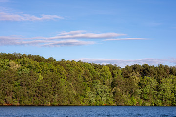 Beautiful landscape, a verdant, green wooded area next to a bright blue lake, with a lovely sky behind, with pink and white fluffy clouds, in upstate South Carolina.