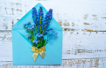  A bouquet of mussari and small blue flowers in a blue envelope is decorated with a golden bow on a light wooden background. postcard. Copy space