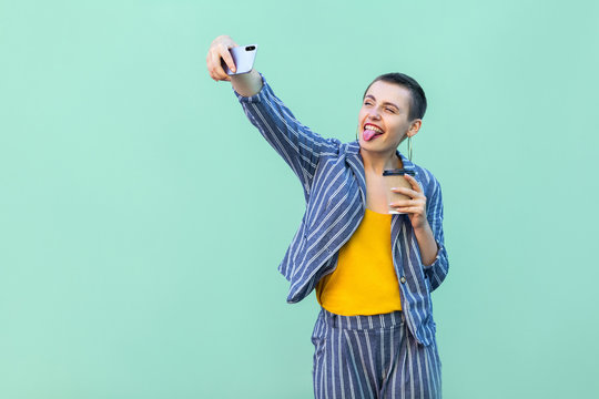 Portrait of beautiful childish blogger with short hair young woman in striped suit standing, making selfie and showing tongue out. Indoor, isolated, studio shot, green background
