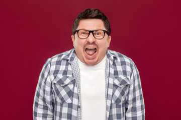 Fototapeta premium Portrait of angry handsome middle aged business man in casual checkered shirt and eyeglasses standing, looking and screaming at camera. indoor studio shot, isolated on dark red background.
