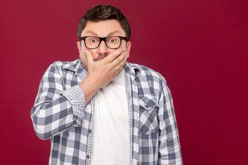 Portrait of shocked middle aged business man in casual checkered shirt and eyeglasses standing, covering his mouth and looking at camera with scared face. studio shot, isolated on dark red background.