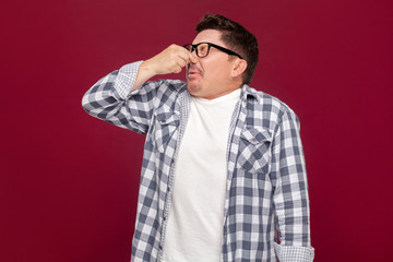 Portrait of bored handsome middle aged business man in casual checkered shirt and eyeglasses standing and blocking his nose because bad smell. indoor studio shot, isolated on dark red background.