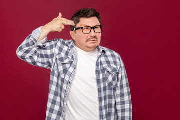 pressure, panics or difficult moments concept. Portrait of alone sad middle aged man in checkered shirt, eyeglasses standing with pistol gesture on head. studio shot, isolated on dark red background.