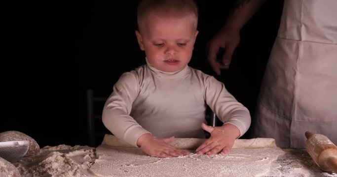 A little boy on a black background helps his father (dad) chef, preparing the dough to roll out for the pizza buns bread cakes and various flour products. Concept of: Little Chef, Baker, Slow motion.