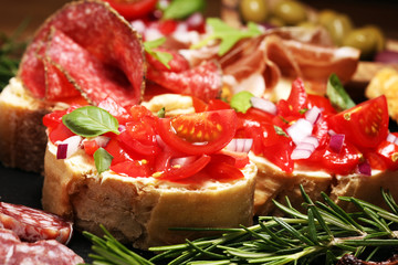Traditional italian antipasto bruschetta appetizer with cherry tomatoes, cream cheese, basil leaves and balsamic vinegar on cutting board with prosciutto, salami, cheese