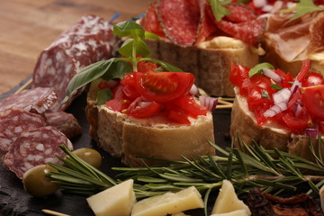 Traditional italian antipasto bruschetta appetizer with cherry tomatoes, cream cheese, basil leaves and balsamic vinegar on cutting board with prosciutto, salami, cheese