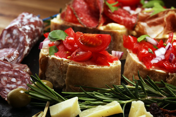 Traditional italian antipasto bruschetta appetizer with cherry tomatoes, cream cheese, basil leaves and balsamic vinegar on cutting board with prosciutto, salami, cheese