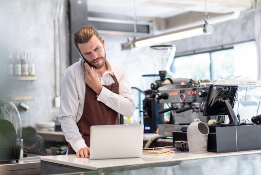 Serious Worried Barista Man Taking A Call On His Mobile Phone  Checking Information Received On His Laptop With A Serious Concerned Expression In Cafe Shop. Startup Owner Small Business Concept.