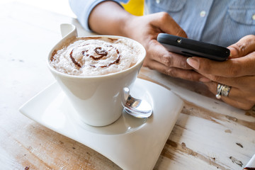 Young woman sitting in coffee shop at wooden table, drinking coffee and using smart phone