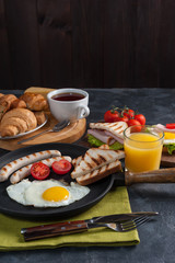 Fried eggs with sausage and vegetables on pan. Breakfast with orange juise and croissant on wooden rustic boards, food background.