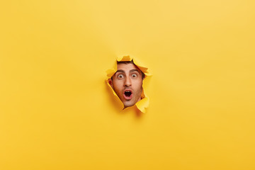 Surprised young man with bristle keeps mouth widely opened, stares through torn paper wall, shows...