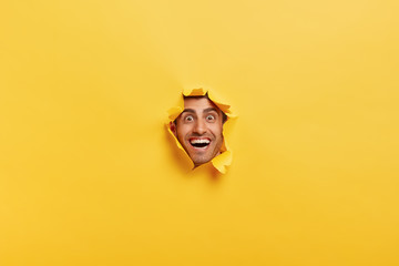 People and happiness concept. Joyous funny young man with thick bristle, looks through torn paper...