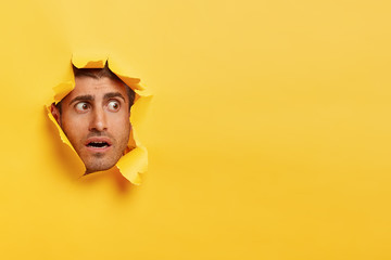 Scared Caucasian man focused aside with surprised facial expression, feels puzzled and nervous, peeks through hole in yellow background, blank copy space for your promotional content or slogan