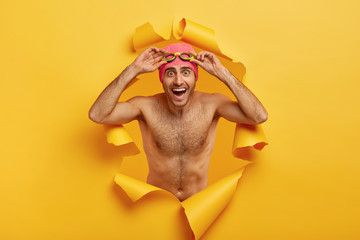 Horizontal shot of happy shirtless man adjustes goggles for swimming, being in good mood, spends wonderful summer holidays with fun, looks through yellow paper wall. People and active rest concept